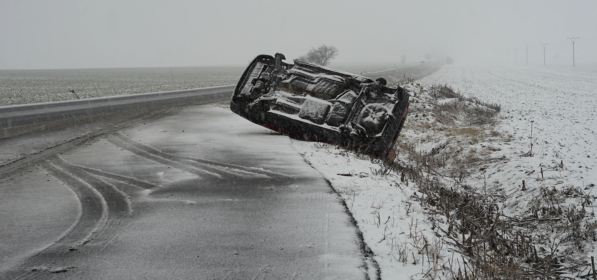 A car is flipped on its side on the side of a snowy road
